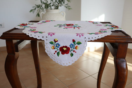 Χειροποίητο τετράγωνο εργόχειρο 45x45 εκ. με πολύχρωμα παραδοσιακά κεντητά λουλούδια σε λευκό βαμβακερό ύφασμα  στεφάνι πάνω σε τραπεζι σε ένα δωμάτιο άδιο  